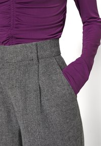 Fialová dlouhá rukávová halenka s nařasenou strukturou na ramenech v kombinaci se šedými kalhotami s herringbone vzorem a vysokým pasem, které mají boční kapsy.