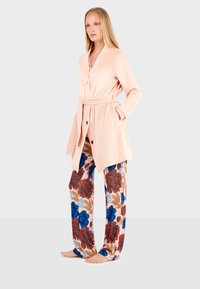 Cardigan lungo rosa chiaro con maniche lunghe, cintura con nodo e bottoni, abbinato a pantaloni larghi con motivo floreale nei colori blu, marrone e crema.