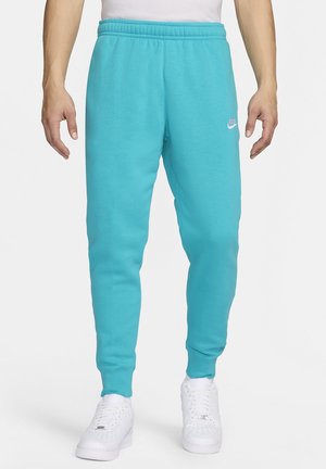 Mężczyzna w jasnoniebieskich spodniach dresowych Nike i białych tenisówkach stoi na jednolitym tle.