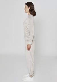 Jasnoszary welurowy dres z długim rękawem, posiadający boczne kieszenie, oraz dopasowane spodnie zwężane ku dołowi. Buty są białe z żółtymi akcentami.