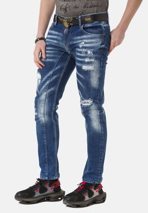 Slim-Fit blaue Jeans aus Denim mit Wisch-Effekten und verwaschenen Details. Trägt einen schwarzen Gürtel mit einer dekorativen Schnalle und schwarze sportliche Schuhe.