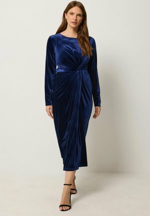 Femme portant une robe midi en velours bleu marine à manches longues avec un détail de nœud torsadé à la taille, associée à des talons noirs à bride cheville.