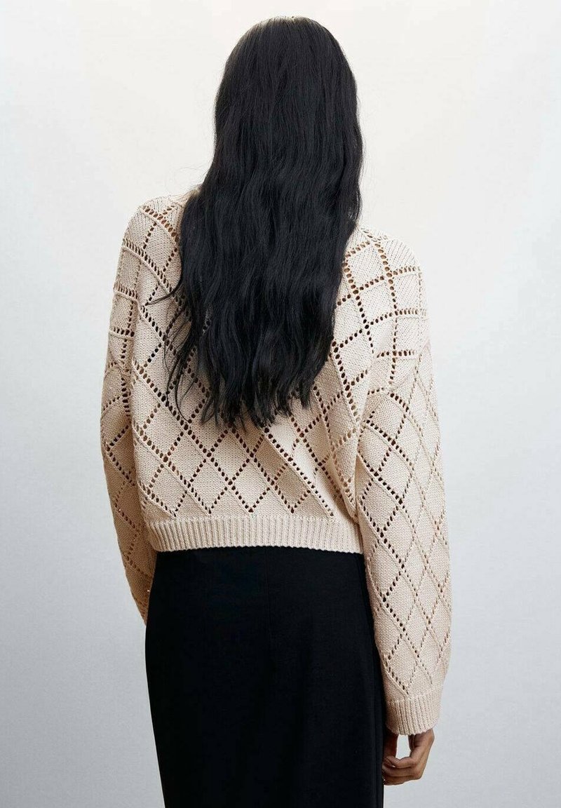 Personne aux longs cheveux noirs portant un pull beige à motif losanges en tricot et un pantalon noir, se tenant face à un mur gris clair uni.