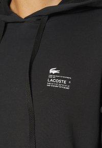 Černá mikina s kapucí, s texturovanými šňůrkami, s bílým logem Lacoste a souřadnicemi tištěnými na přední straně. Vyrobena z hladkého materiálu ze směsi bavlny.