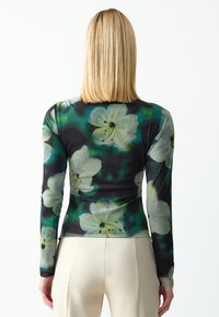 Haut à manches longues en tissu transparent avec un motif floral vert et noir agrémenté de fleurs blanches. Texture lisse et design ajusté.