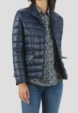 Kvinde iført en marineblå quiltet jakke over en sort og hvid blomstret skjorte med blå jeans mod en ensfarvet baggrund.