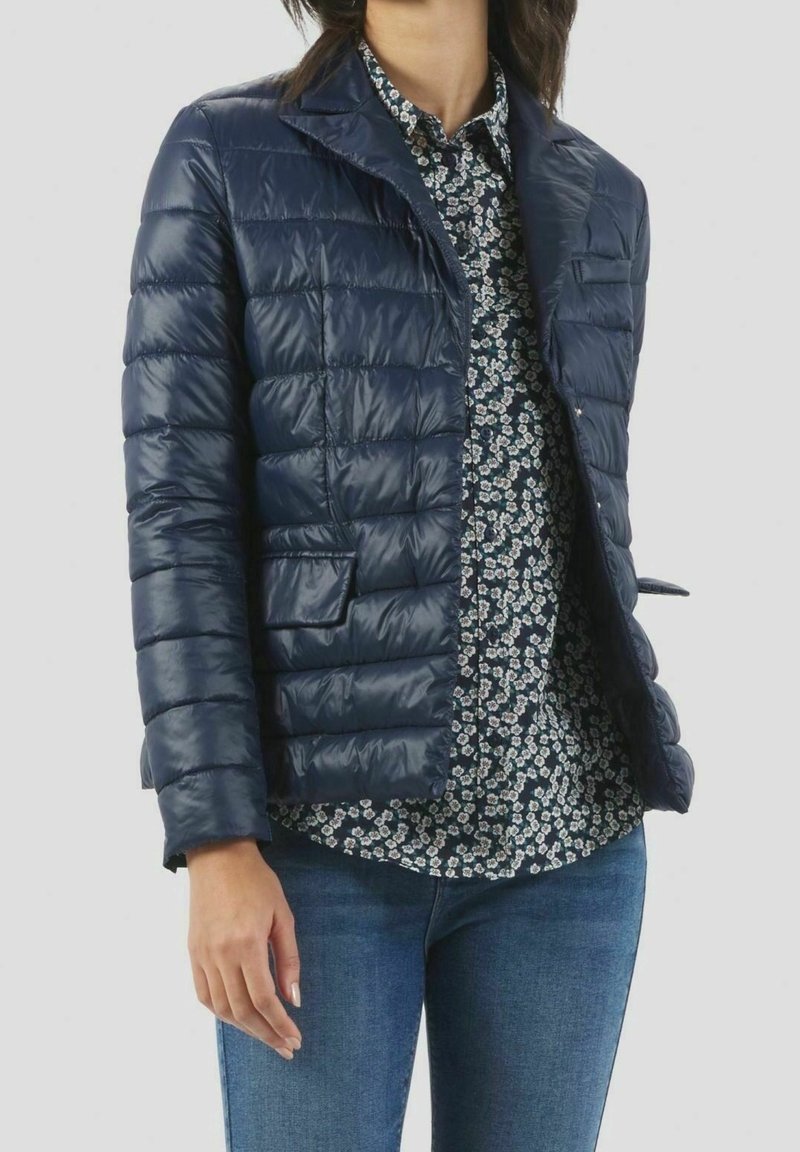 Femme portant une veste matelassée bleu marine par-dessus une chemise fleurie noire et blanche avec un jean bleu sur un fond uni.