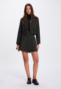 Giacca corta a quadri bianco e nero con tasca frontale, abbinata a una gonna coordinata con motivo a diamante e mocassini neri.