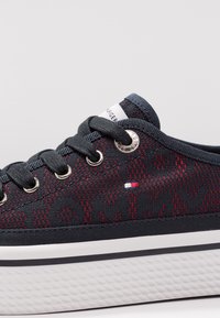 Schwarze und rote Sneaker mit einem gemusterten Textilobermaterial, silbernen Ösen und einer weißen strukturierten Außensohle. Verfügt über ein markiertes Etikett auf der Zunge.