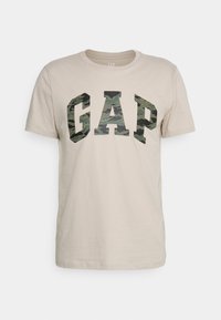 Camiseta de algodón beige con un gran logo "GAP" en camuflaje de color verde oscuro y negro en el centro. Mangas cortas y diseño de cuello redondo.