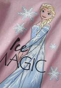 Un tissu rose clair présentant un graphique d'un personnage en robe bleue avec des flocons de neige, le texte "Magie de Glace" en noir, et des accents pailletés.