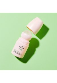 Rullande deodorant i en ljusrosa plastbehållare med ett rundat applikatorlock och grön text, placerad mot en mintgrön bakgrund.