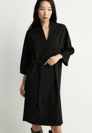 Woman wearing a black knee-length dress with wide sleeves and a waist tie, standing against a plain light background.