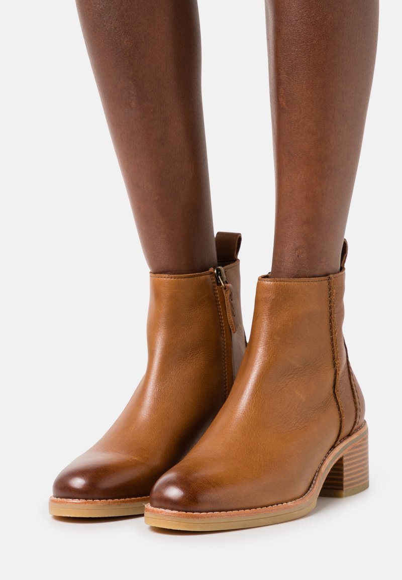 Botas de cuero marrón hasta el tobillo con puntas redondeadas, cremalleras laterales, un tacón apilado y una textura suave. Con acentos de costura y una suela ligera.