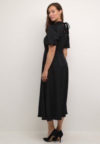Rochie midi neagră dintr-un material neted, cu mâneci bufante și detaliu legat la spate. Asortată cu pantofi cu toc înalt negri.
