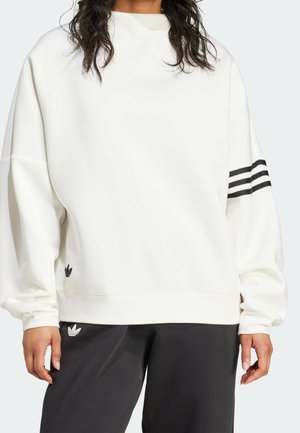 Person trägt ein weißes Sweatshirt mit schwarzen dreifachen Streifen am linken Ärmel und schwarze Hose, beide mit Adidas-Logos.
