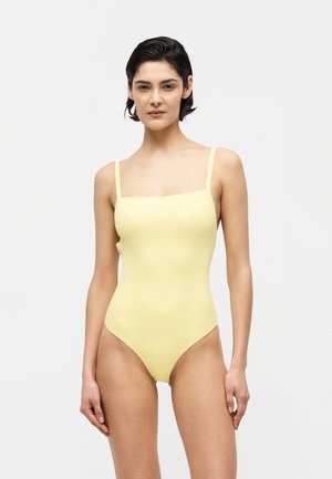 Žena stojí a má na sebe jednoduché svetložlté jednodielne plavky s tenkými ramienkami na bielom pozadí.