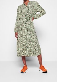 Abito midi verde con motivo, vita con cinturino, maniche lunghe e chiusura con bottoni. Indossato con sneakers arancioni per creare un contrasto.