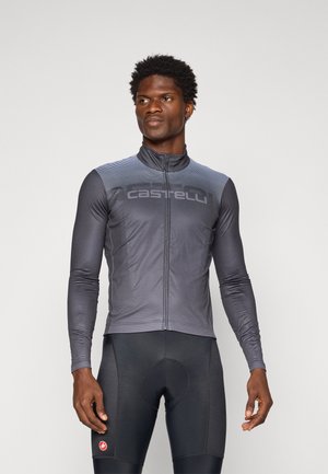 Ein Mann trägt ein dunkelgraues Langarm-Radsporttrikot und schwarze gepolsterte Radsportshorts und steht vor einem schlichten Hintergrund.