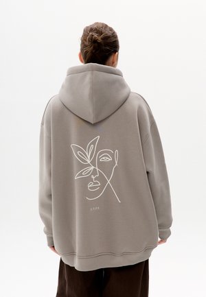 Personne portant un sweat à capuche gris surdimensionné avec un dessin minimaliste en ligne blanche d'un visage et des feuilles au dos, debout devant un fond uni.