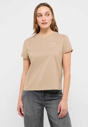 Frau trägt ein beigefarbenes T-Shirt mit kurzen Ärmeln und dem Schriftzug "Mustang Jeans" auf der Brust sowie ausgewaschene schwarze Jeans, steht vor einem einfarbigen Hintergrund.