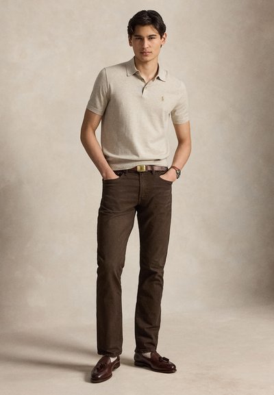 Νεαρός άνδρας στέκεται με τα χέρια στις τσέπες, φοράει μπεζ πόλο μπλούζα, σκουρόκαφέ παντελόνι, καφέ ζώνη, ρολόι και καφέ μοκασίνια με φούντες.