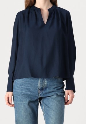 Personne portant un chemisier bleu marine à manches longues avec une encolure fendue et un jean en denim bleu clair, debout devant un fond uni.