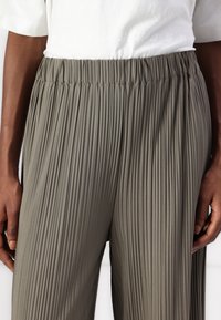 Gros plan sur une personne portant un pantalon plissé ample vert olive avec une taille élastique et un haut court à manches courtes texturé blanc.