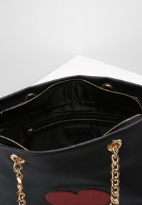 Svart läder tote-väska med ett rött hjärtmotiv, guldkedjehandtag och ett blixtlåsfack. Insidan har en slät, fodrad tyg.