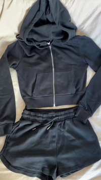 Felpa nera con zip, tasche frontali e cappuccio foderato, abbinata a pantaloni corti neri coordinati con cintura elastica e lacci.