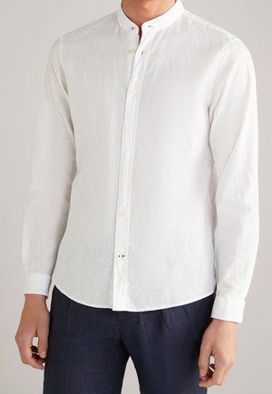 Homme portant une chemise blanche à manches longues avec col mandarin et boutons, associée à un pantalon bleu marine foncé.