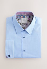 Chemise boutonnée bleu clair avec un motif texturé, présentant un imprimé floral sous le col et un détail de poignet à un seul bouton.