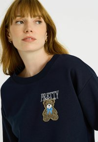 Sudadera azul marino con cuello redondo, con un oso marrón bordado que lleva una pajarita azul. La palabra "PRETTY" está cosida encima del oso en color blanco.
