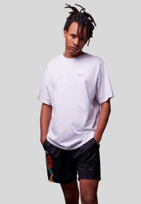 Weißes T-Shirt mit kurzen Ärmeln und Logo auf der Brust, kombiniert mit schwarzen Shorts, die ein buntes Papageienmotiv und einen strukturierten Bund aufweisen.