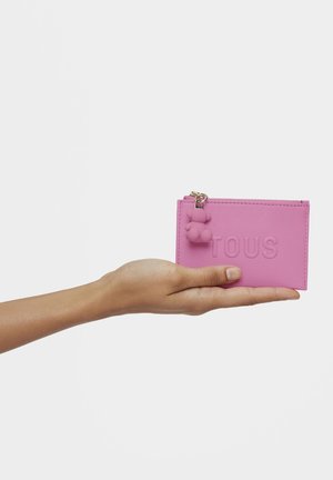Portacarte in pelle rosa con la scritta "TOUS" in rilievo, caratterizzato da un piccolo ciondolo a forma di orsetto e chiusura con zip. Tenuto in mano per riferimento alle dimensioni.