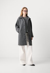 Grå struktureret frakke med krave, knapper foran, sidelommer, båret over en ribbet grå sweater og hvide bukser med vide ben samt sorte sko.