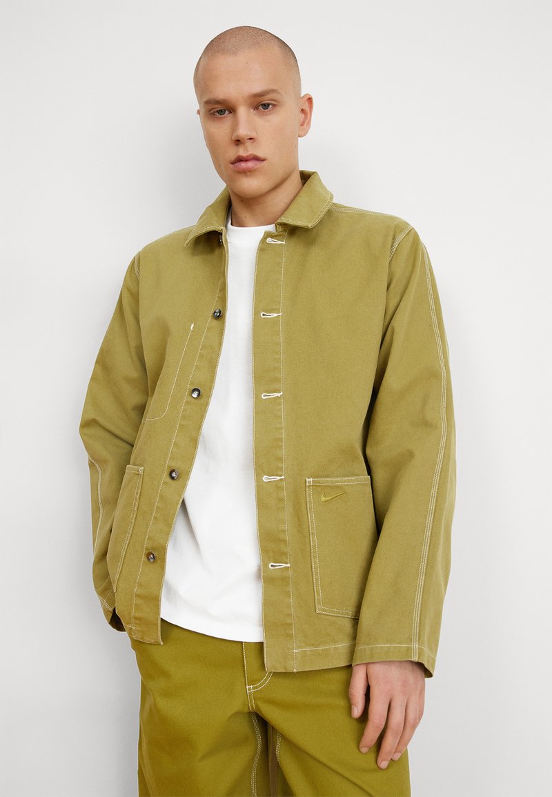 Chaqueta de lona verde olivo con costuras en contraste, botones a presión y un bolsillo lateral con un sutil logo. Llevada sobre una camiseta blanca.
