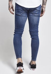 Jeans de denim azul de corte ajustado con deslavado y dos bolsillos traseros. Combinados con zapatillas Vans negras con suela blanca y logotipo rojo.