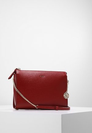 Sac bandoulière DKNY en cuir rouge avec une chaîne dorée et un pendentif rond portant la marque, présenté sur un socle blanc sur un fond uni.