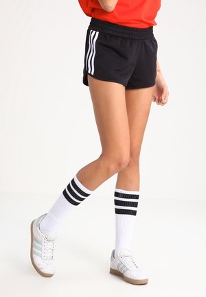 Personne portant un short de sport noir avec des bandes blanches, des chaussettes blanches montantes avec des bandes noires et des baskets blanches, debout sur un fond blanc.