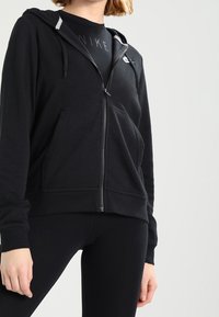 Person wearing black zip-up hoodie over black Nike shirt and black leggings, standing against plain light background.