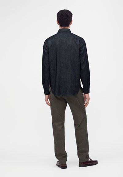 Homme debout de dos, portant une chemise à manches longues sombre, un pantalon vert olive et des chaussures marron sur un fond blanc uni.