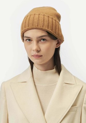 Tan knitted beanie with ribbed texture, featuring a rolled cuff. Worn with a beige high-neck sweater and cream-colored coat.