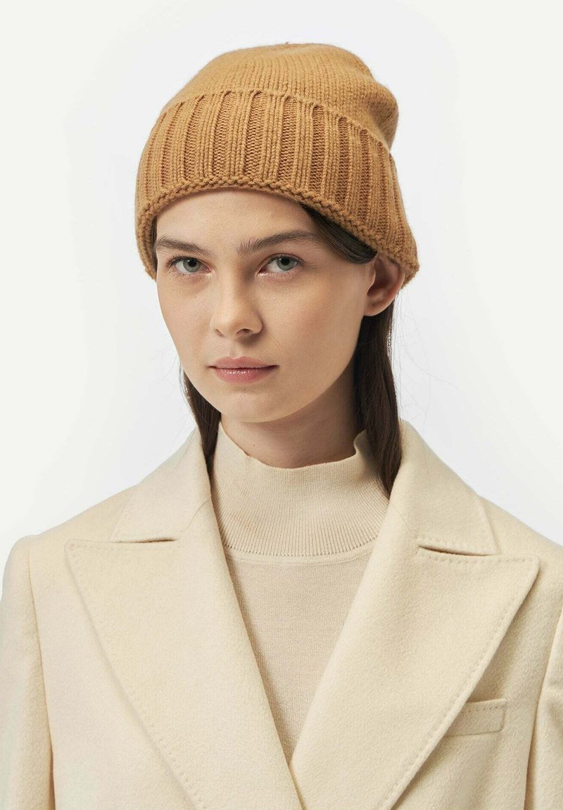Bonnet en tricot couleur sable avec une texture côtelée, doté d'un revers. Porté avec un pull à col montant beige et un manteau couleur crème.