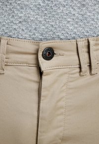 Pantalones de algodón beige con cierre de botón que presenta una etiqueta negra. La tela tiene una textura suave y detalles de costura visibles.