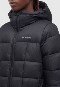 Човек, носещ черно пухено яке Columbia с качулка, предна цип и видимо лого на гърдите.