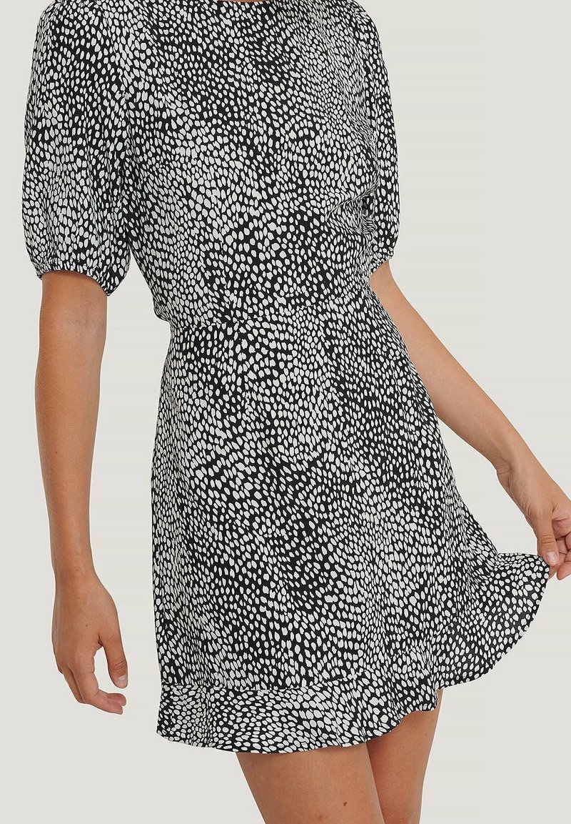 Femme portant une robe courte noire et blanche à pois avec des manches bouffantes, tenant l'ourlet de la robe sur un fond uni.