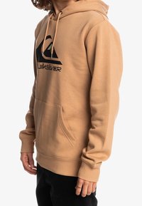 Hoodie beige realizzato in morbido cotone misto con un grande logo nero e un triangolo di accentuazione verde. Presenta una tasca frontale e cappuccio con coulisse.