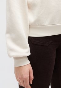 Εκρού φούτερ με ριμπ τελείωμα και μανσέτες, εμφανιζόμενο με παντελόνι από κορντόρι σκούρου καφέ. Ομαλή υφή και ελαφρώς φαρδιά σχεδίαση στα μανίκια.