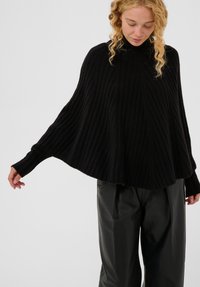 Moteris su garbanotais šviesiais plaukais, apsivilkusi juodą raukšlių megztinį su plačiomis rankovėmis ir juodomis odinėmis kelniomis, žiūri žemyn.